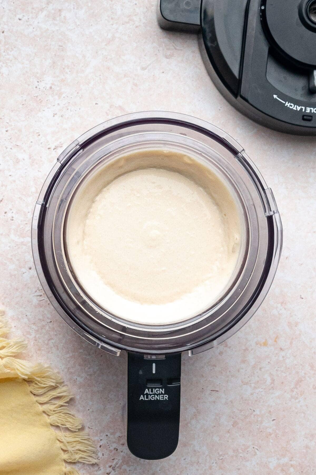 A food processor filled with a smooth, creamy mixture sits on a light-colored countertop next to its detached lid and a yellow cloth.