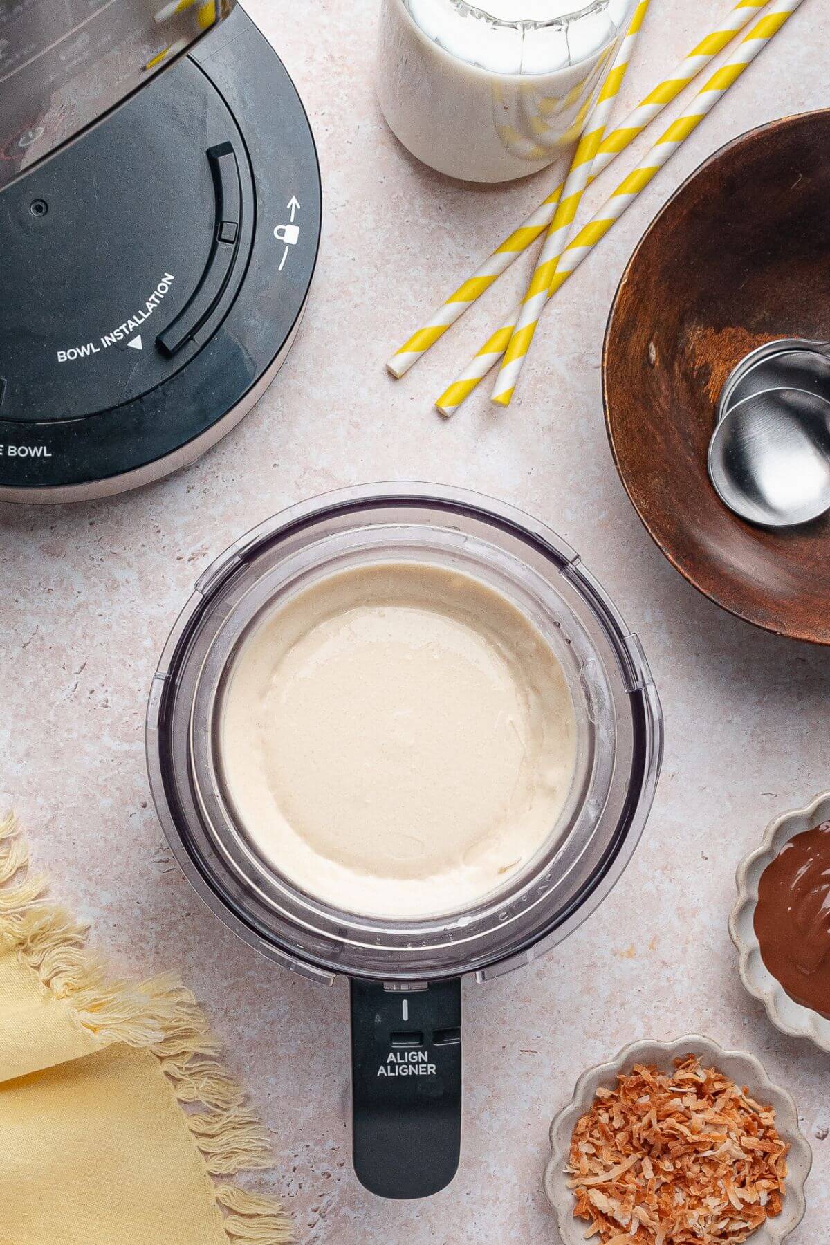 Top-down view of a food processor with a creamy mixture inside, surrounded by a blender base, a bowl of spoons, striped straws, toasted coconut, and a dish of melted chocolate.