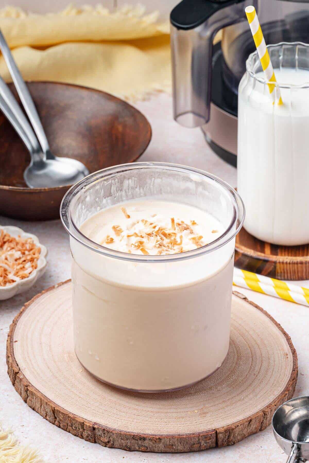A glass container of creamy dessert topped with toasted coconut sits on a wooden coaster, with a jar of milk and kitchen utensils in the background.