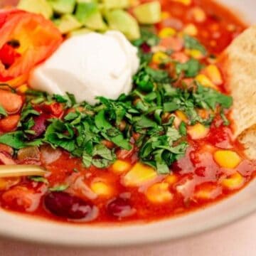 A bowl of chili with beans, corn, avocado, sour cream, chopped cilantro, a sliced pepper, and tortilla chips on the side.