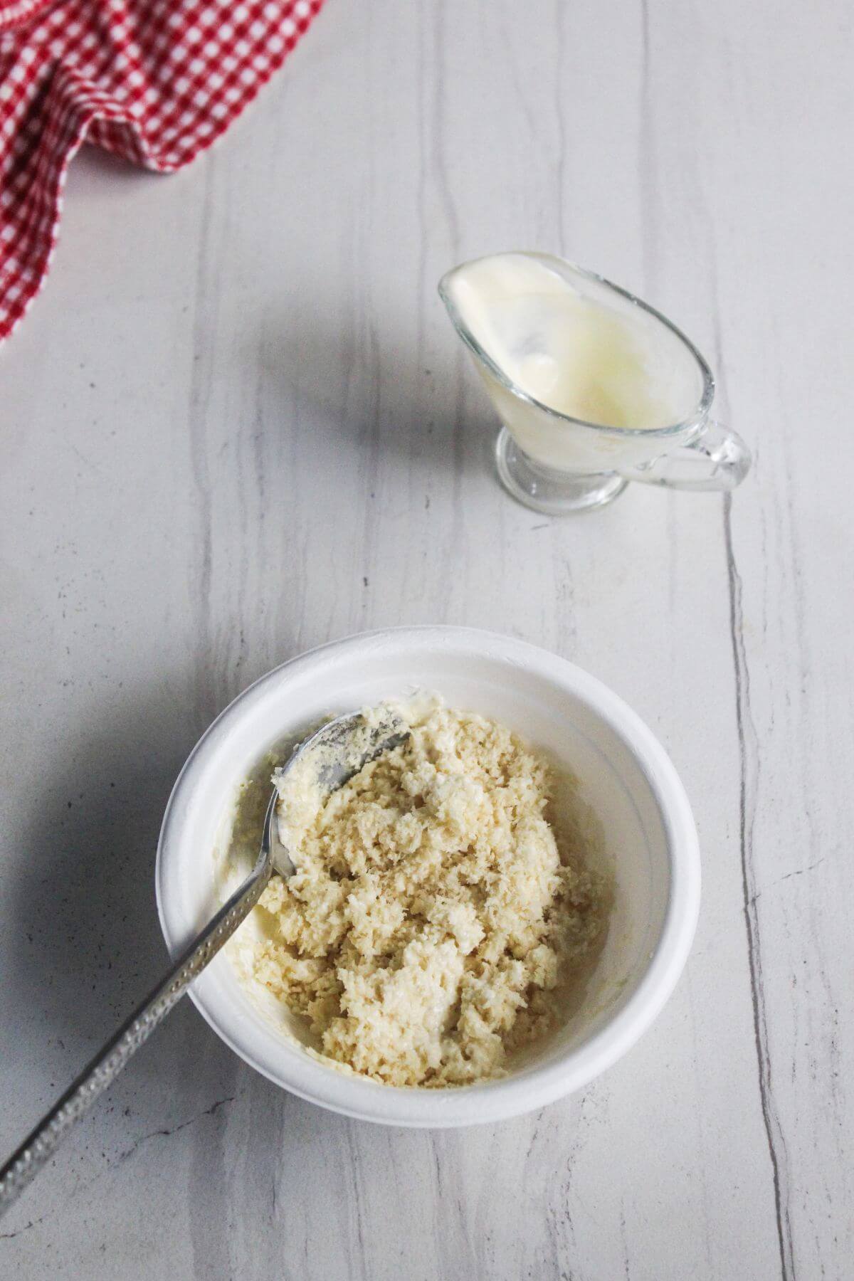 A white bowl of breadcrumb mixture with a spoon beside a small glass gravy boat of white liquid on a light countertop.