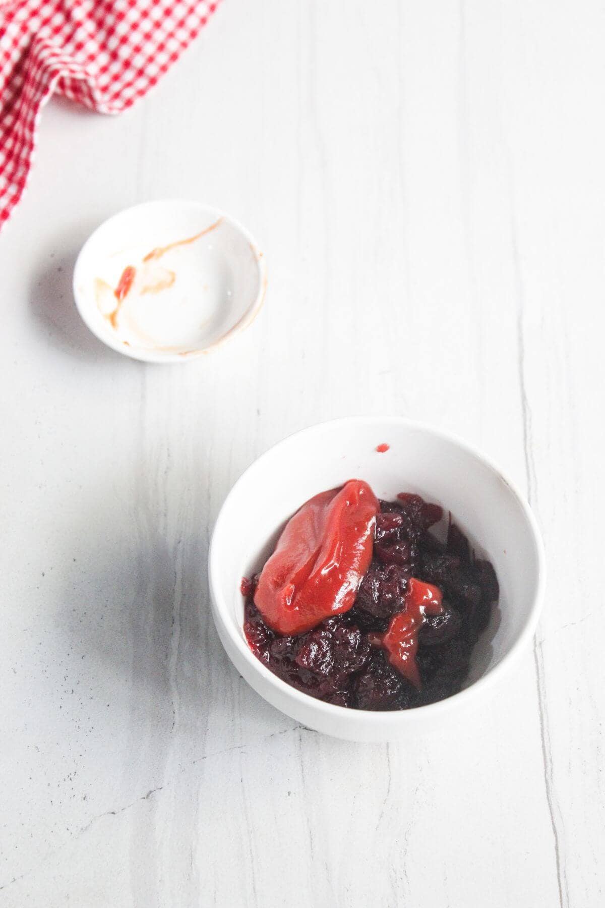 A white bowl with dark stewed fruit and red sauce sits on a white surface; a small empty dish and a red-checkered cloth are nearby.