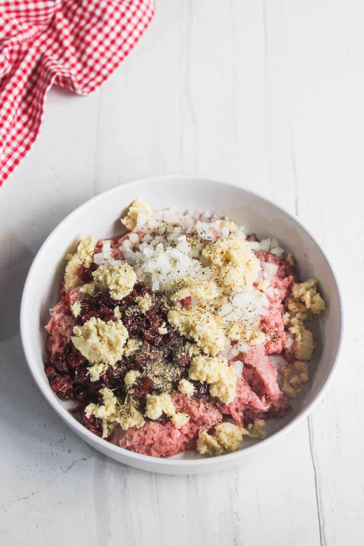 A white bowl filled with ground meat, chopped onions, breadcrumbs, seasonings, and dried cranberries, on a white surface next to a red and white checkered cloth.