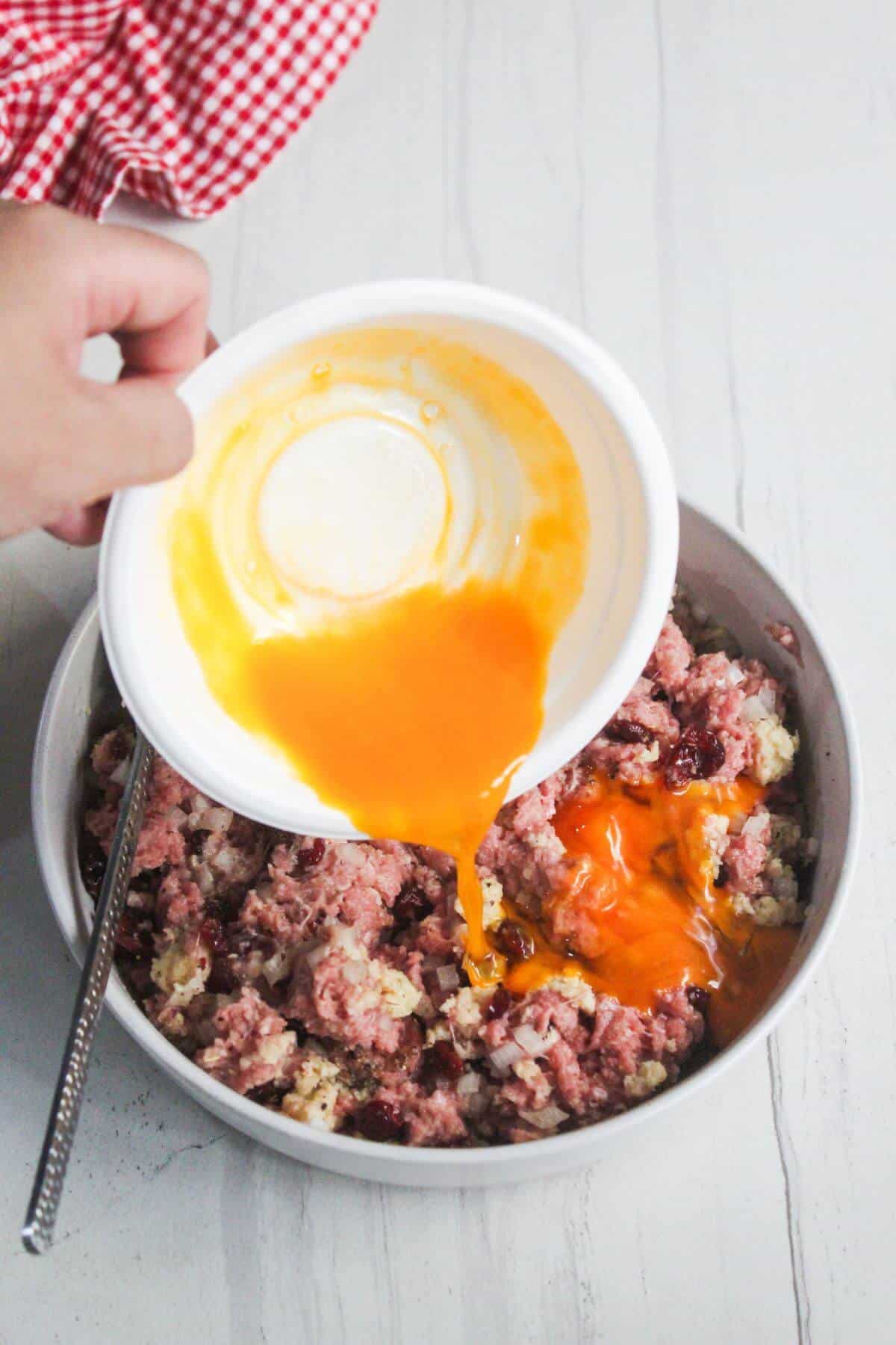 A hand pours beaten eggs from a white bowl into a mixture of ground meat and other ingredients in a larger bowl on a light surface.