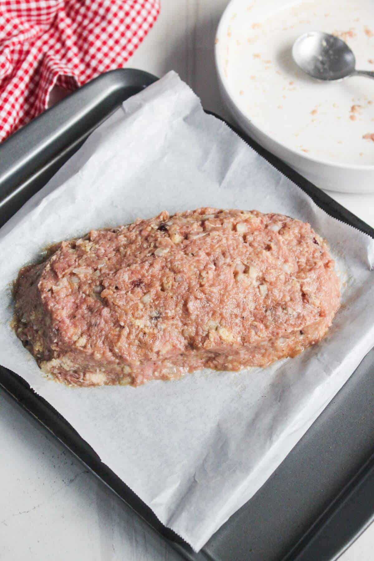 Uncooked meatloaf mixture shaped into a loaf sits on a parchment-lined baking tray next to a spoon and plate.