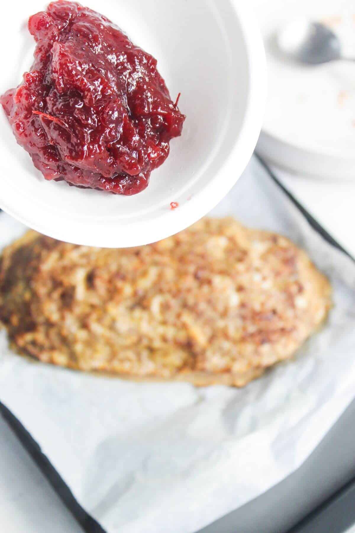A white bowl with cranberry sauce held above a baked meatloaf on a parchment-lined baking sheet.