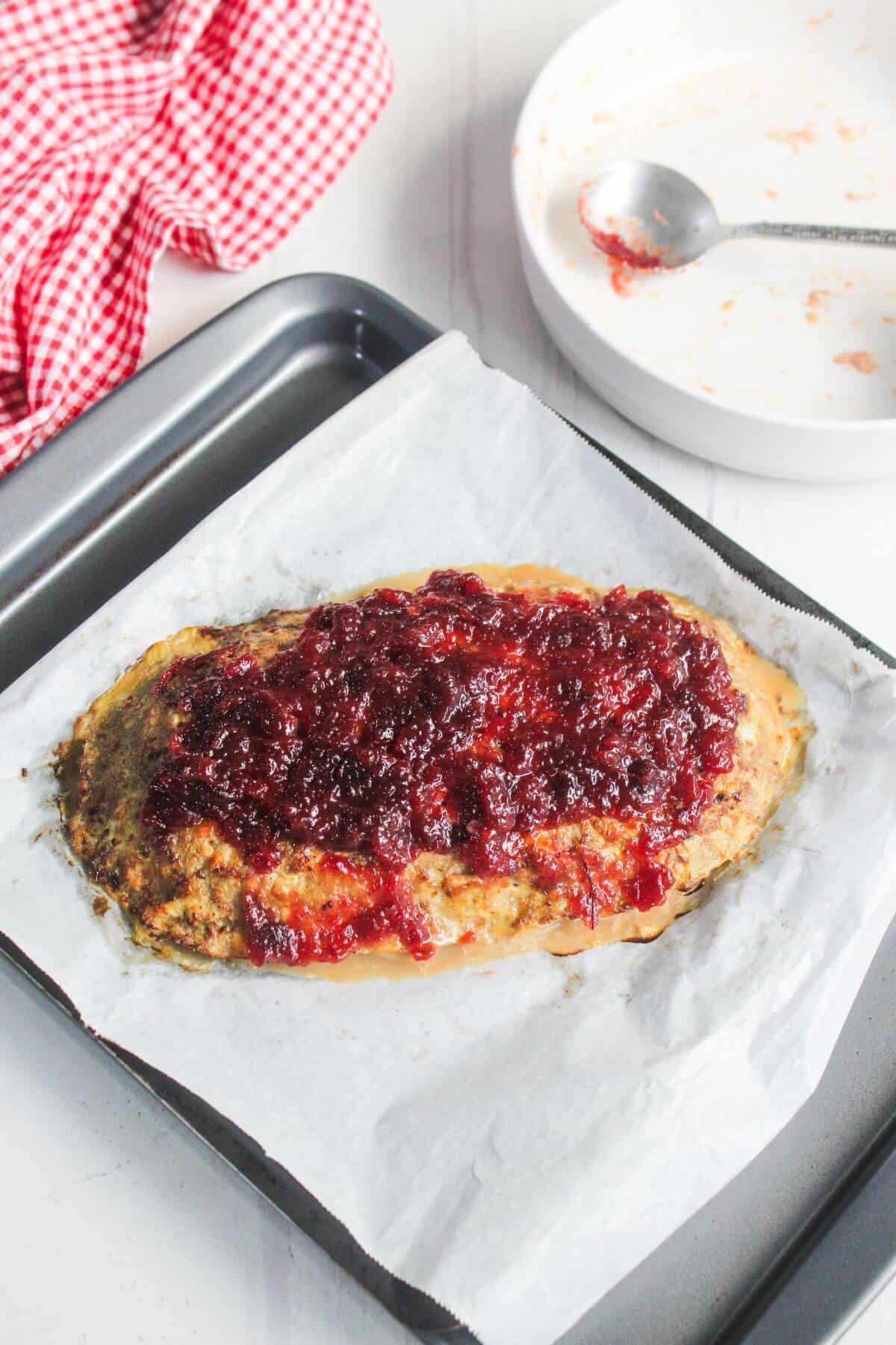A baked meatloaf topped with red sauce sits on parchment paper in a baking tray, with a used plate and spoon in the background.