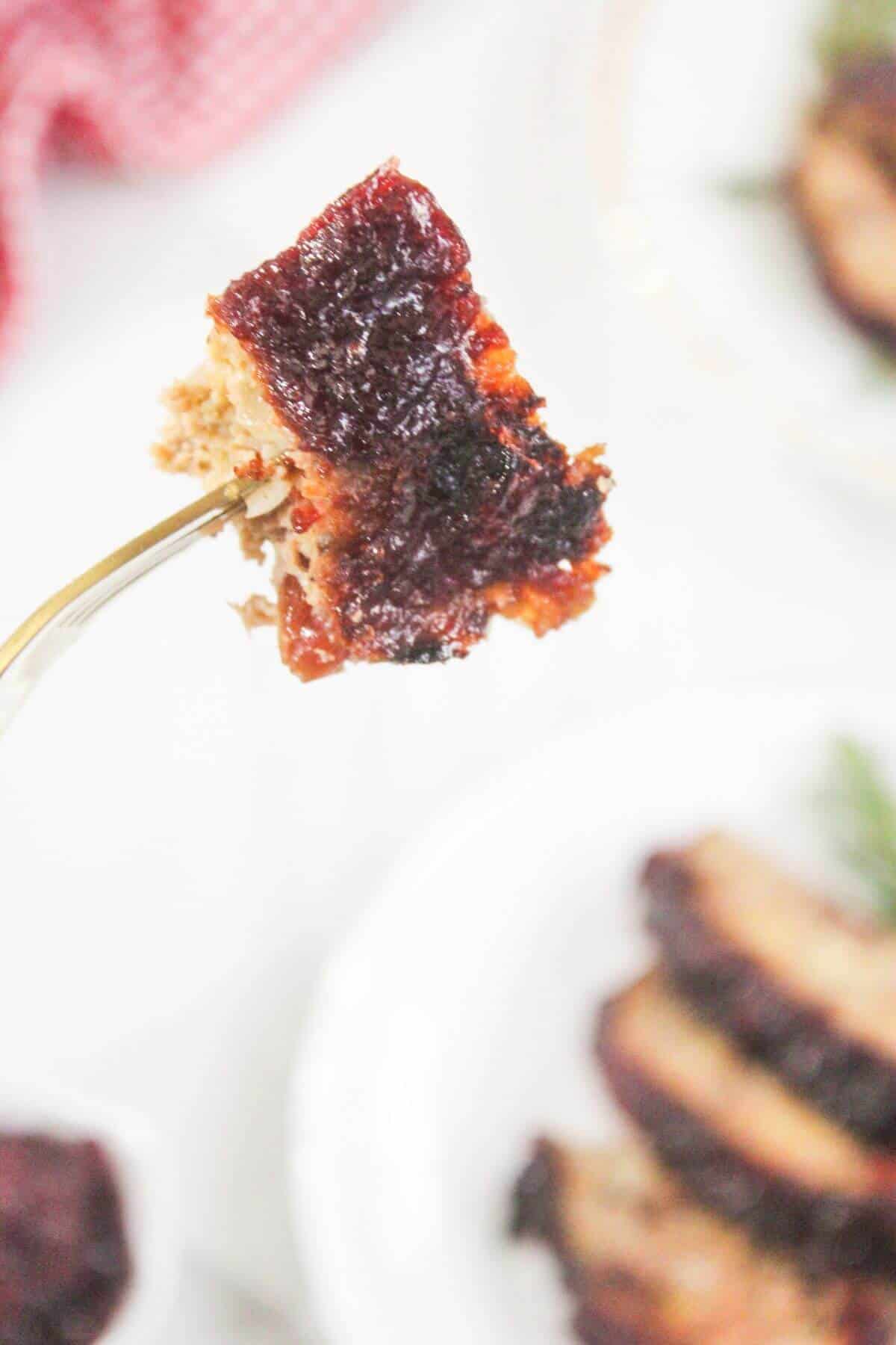 A fork holds a piece of meatloaf topped with a layer of dark red glaze; more slices are visible on a white plate in the background.