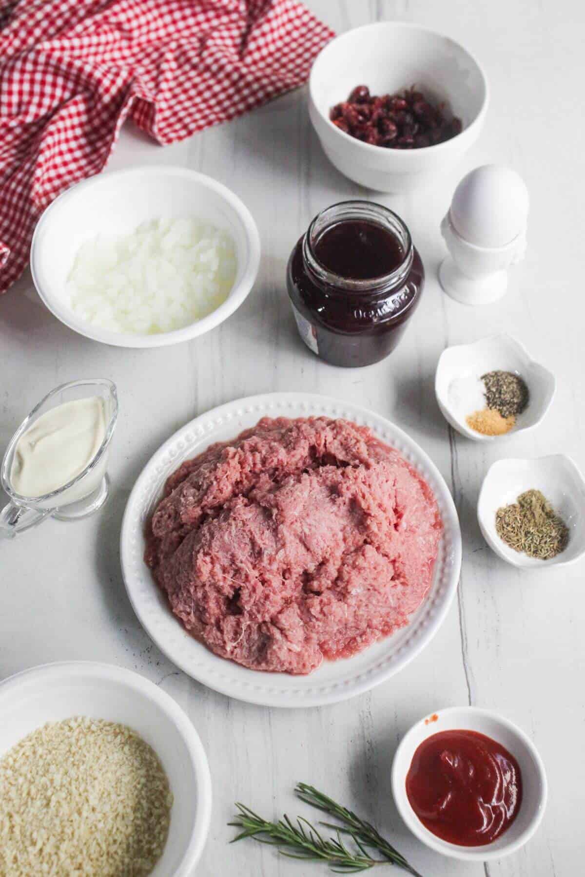Ground meat on a plate surrounded by bowls of chopped onions, breadcrumbs, herbs, seasonings, ketchup, cream, cranberries, a jar of jam, an egg, and a sprig of rosemary on a white surface.
