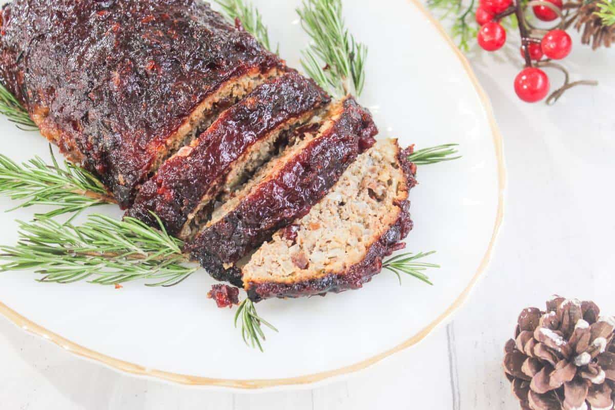 Sliced meatloaf with a dark glaze arranged on a white plate, garnished with rosemary sprigs; pinecones and red berries nearby.