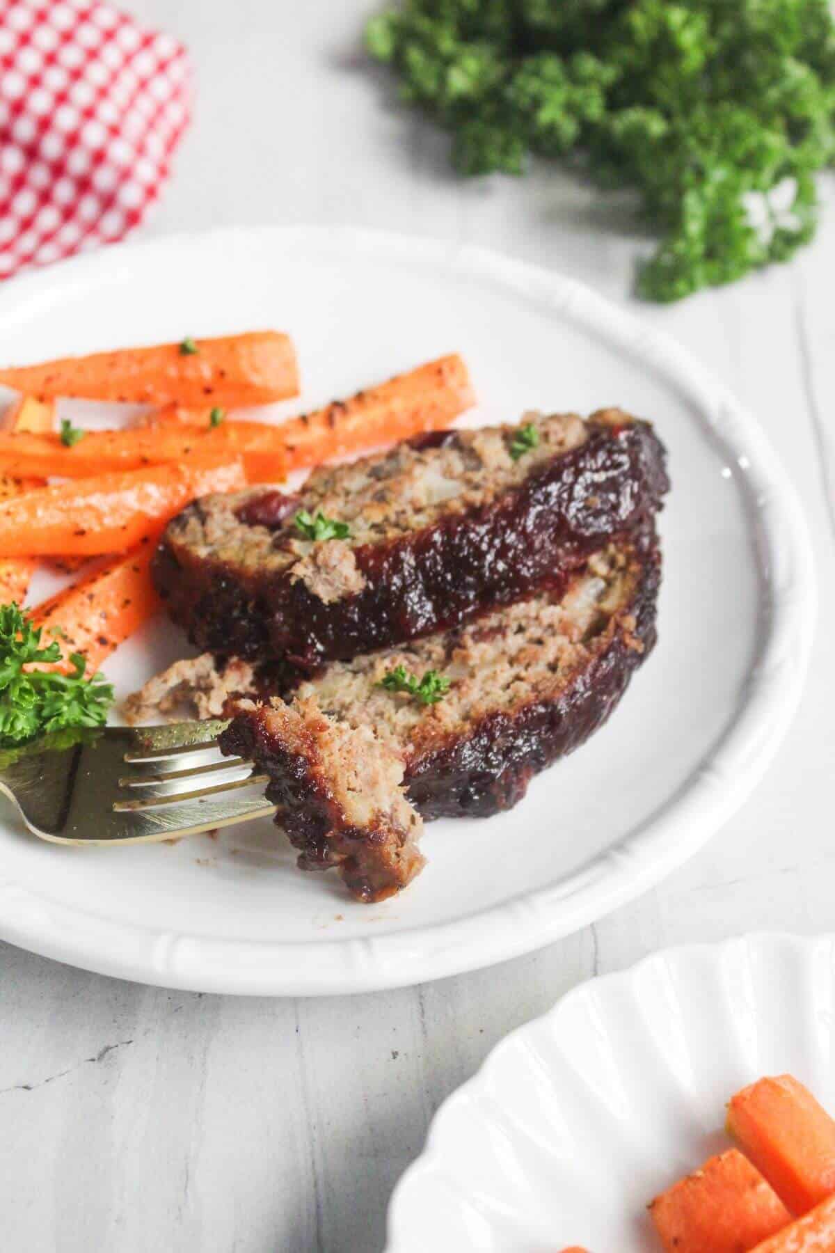 A white plate with two slices of glazed meatloaf, roasted carrots, and a fork holding a piece of meatloaf. Parsley garnish is on the side.