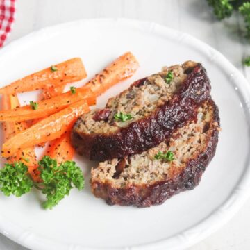 Two slices of glazed meatloaf and roasted carrot sticks are served on a white plate, garnished with fresh parsley.