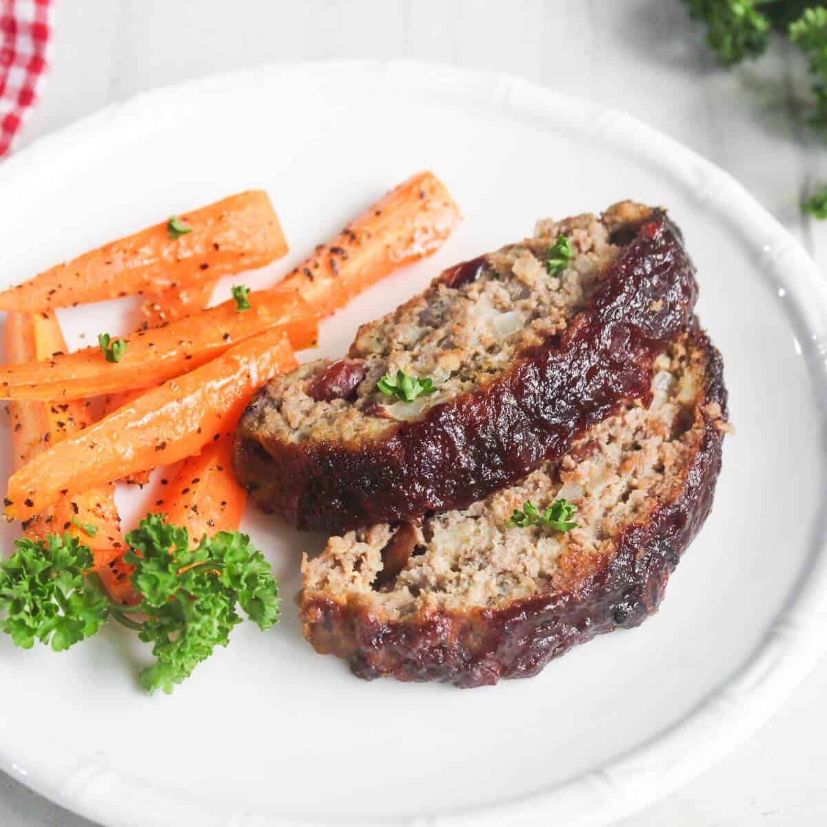 Two slices of glazed meatloaf and roasted carrot sticks are served on a white plate, garnished with fresh parsley.