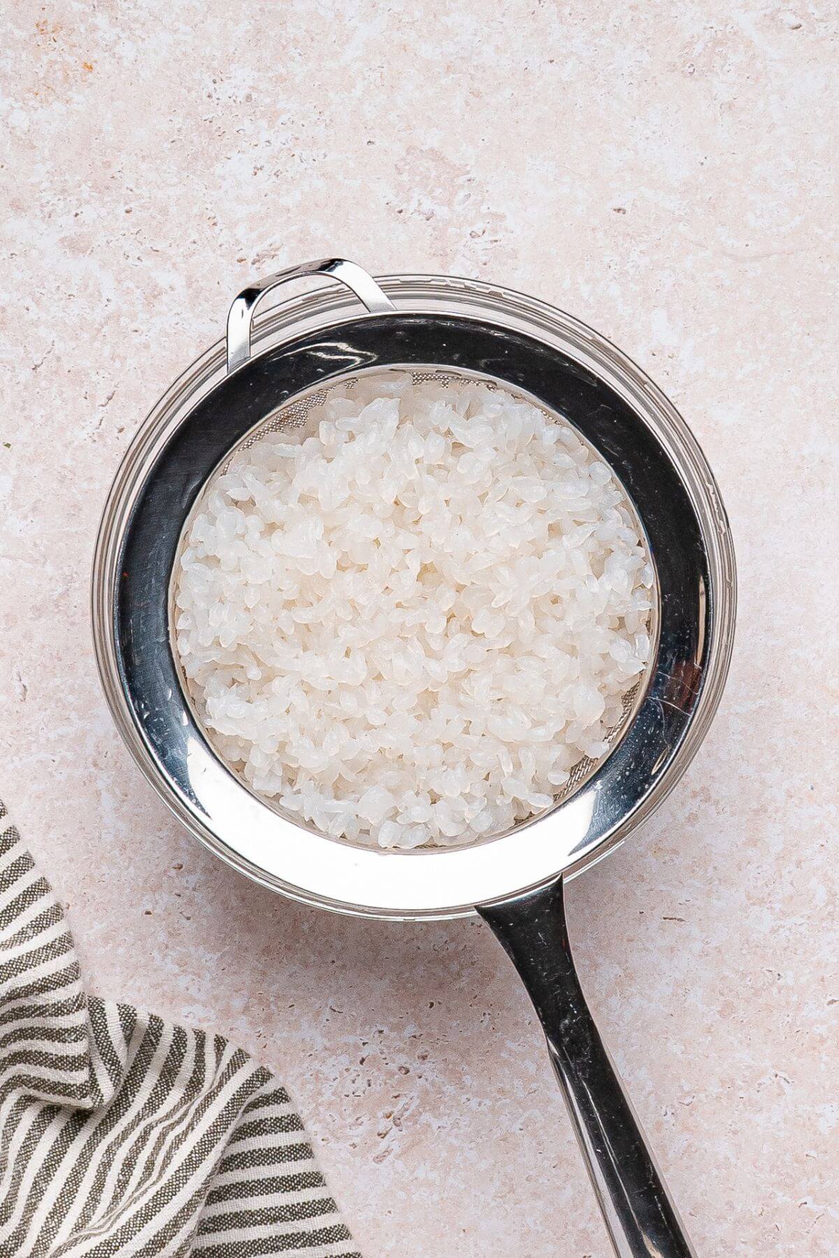 A metal strainer filled with white rice sits on a light-colored countertop next to a striped cloth.