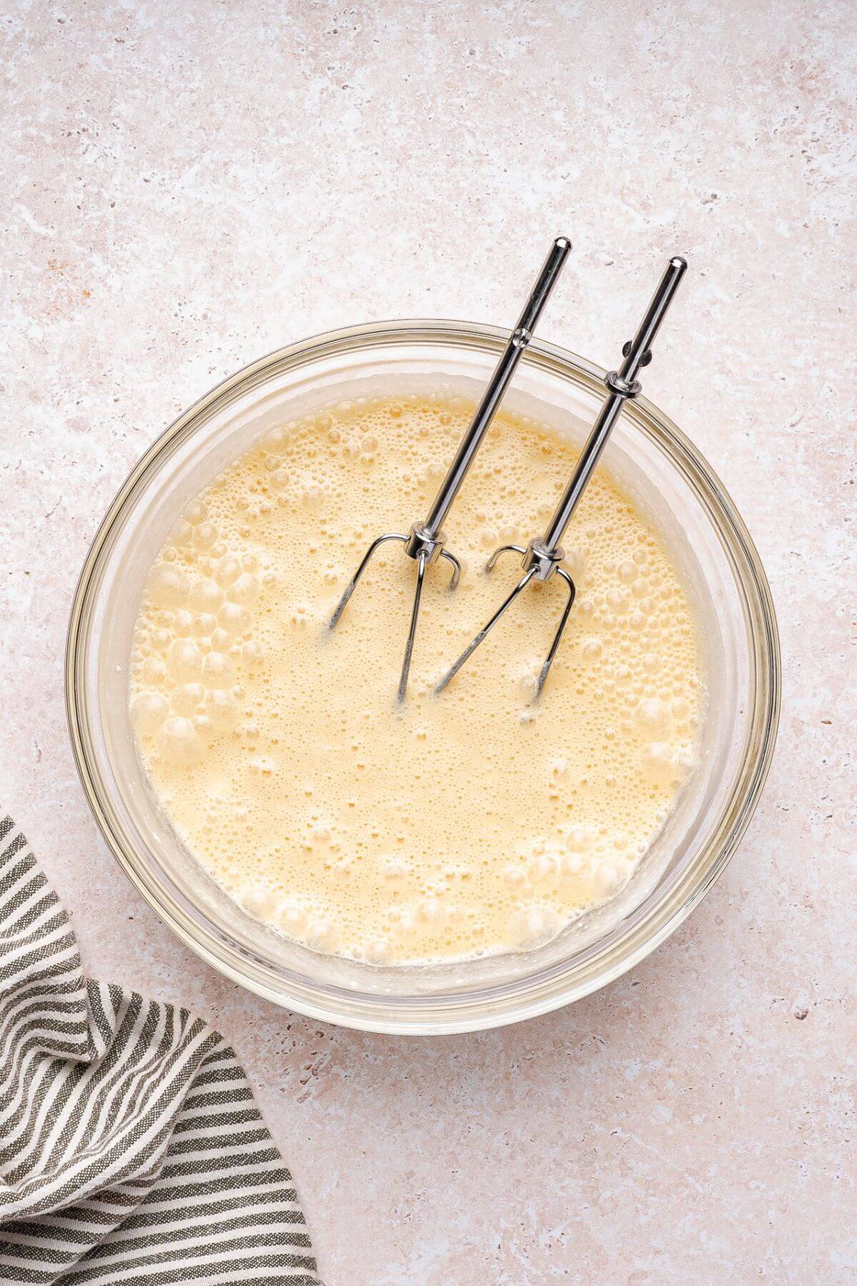 A glass bowl filled with beaten eggs and a pair of metal beaters resting inside, on a light countertop next to a striped cloth.