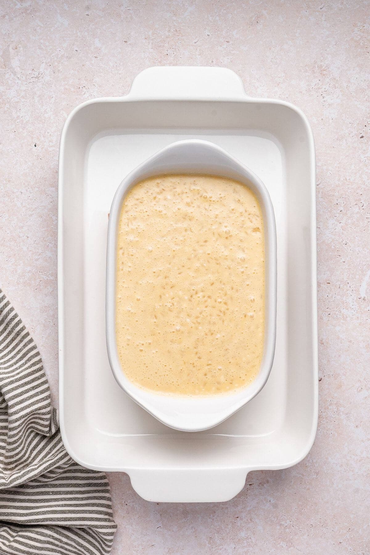 A white baking dish filled with a light yellow, bubbly batter, placed inside a larger white pan on a speckled countertop next to a striped cloth.