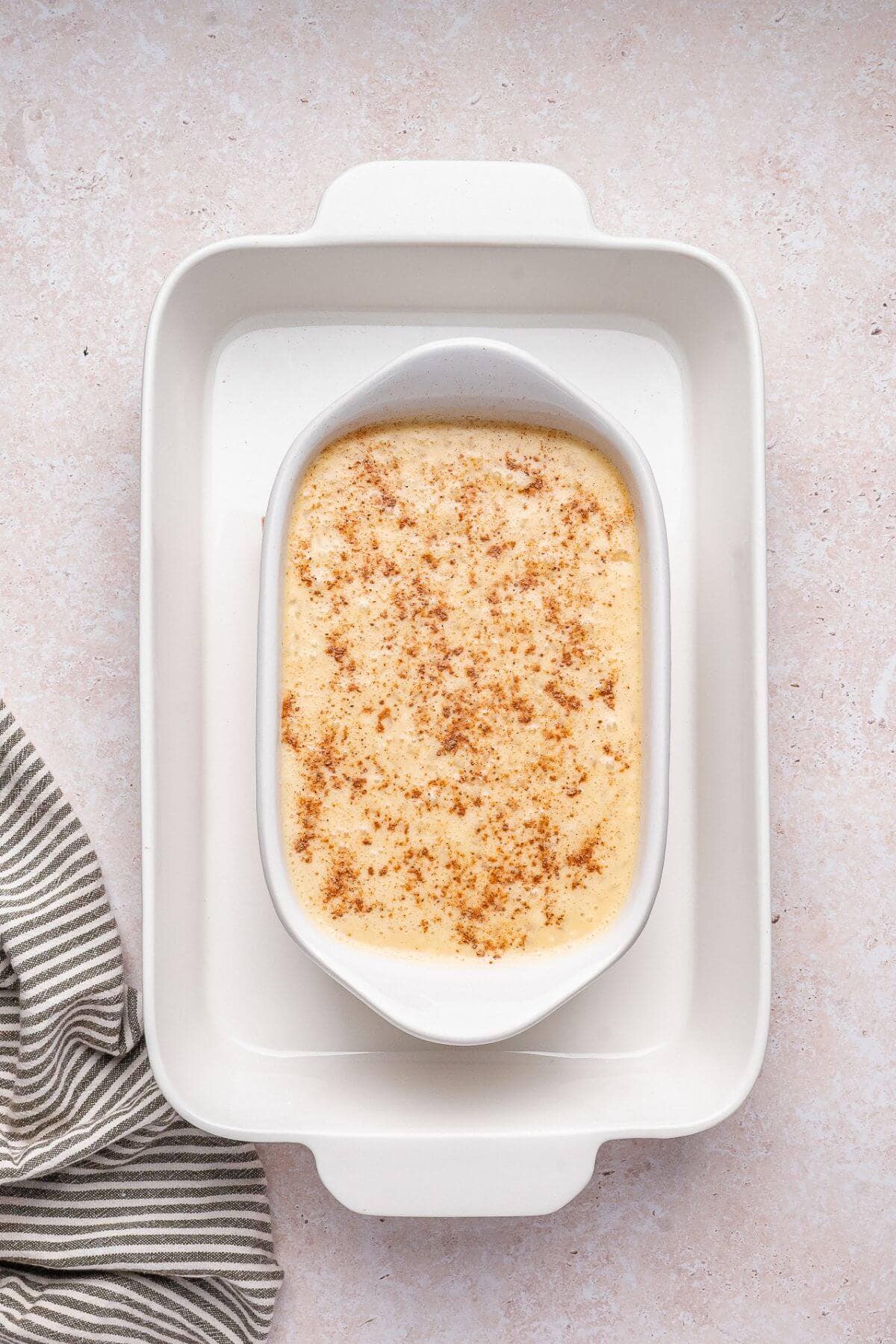 A rectangular dish of baked custard topped with ground spices sits inside a larger white baking dish on a light countertop, next to a striped cloth.