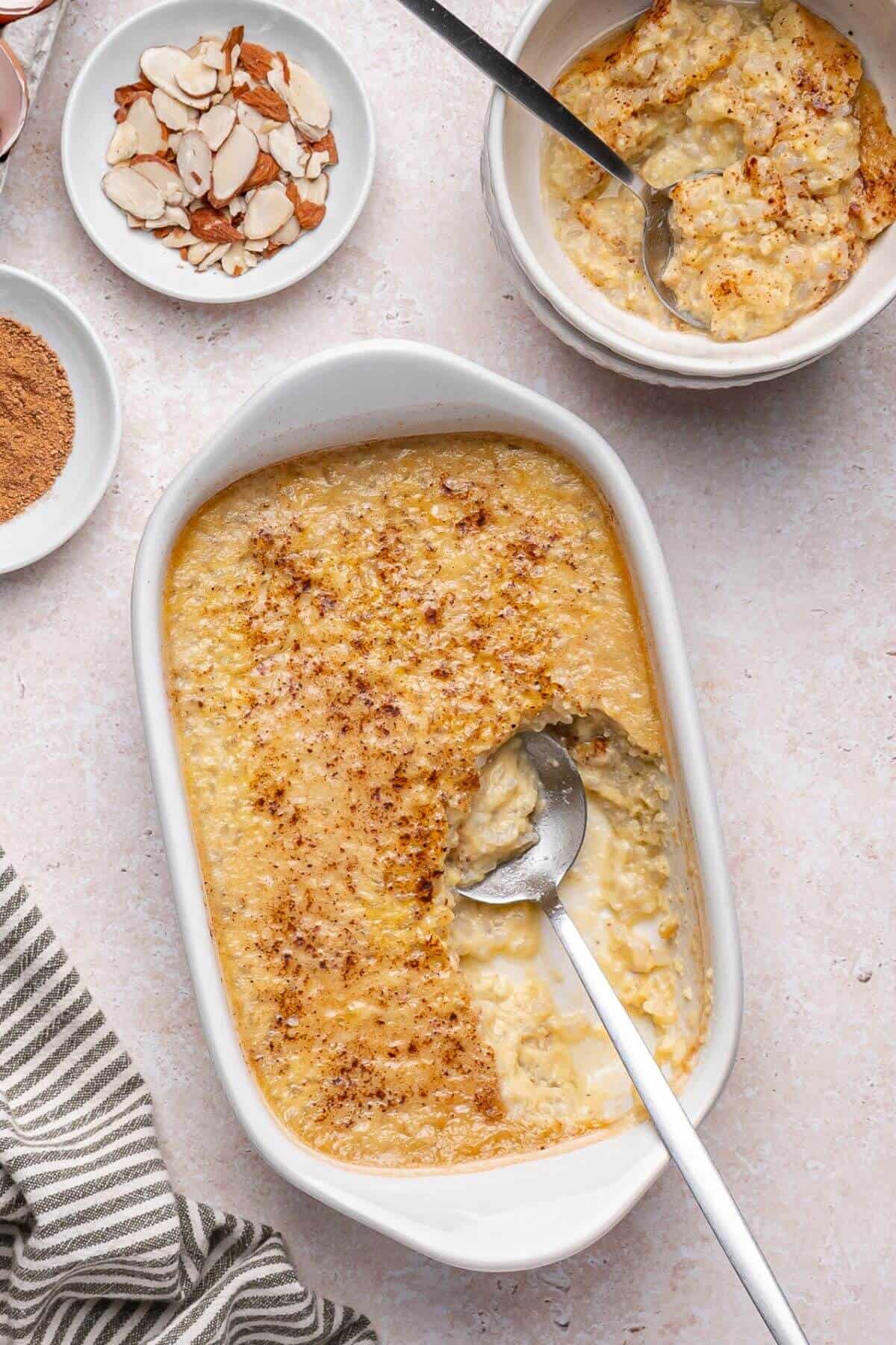 A white baking dish of baked rice pudding with a serving spoon, next to a bowl of pudding, sliced almonds, and a plate of cinnamon on a light surface.