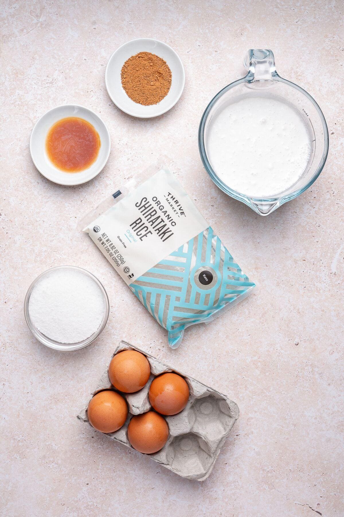 Six ingredients are arranged on a light surface: eggs in a carton, coconut milk in a jug, shirataki rice, granulated sweetener, vanilla extract, and ground cinnamon in small bowls.
