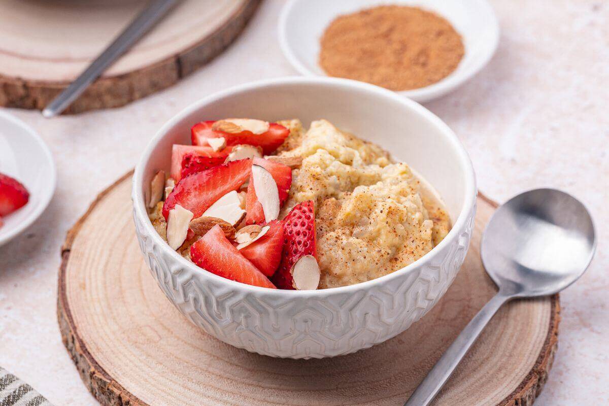 A bowl of oatmeal topped with sliced strawberries and almonds sits on a wooden coaster next to a spoon and a small plate of brown sugar.