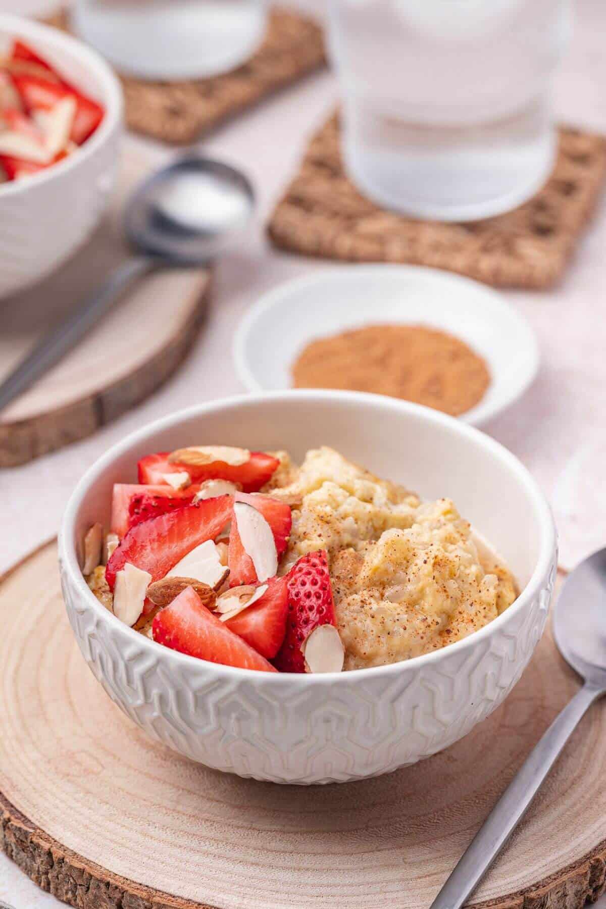 A bowl of oatmeal topped with sliced strawberries and almonds sits on a wooden coaster, with a spoon and a small dish of cinnamon in the background.