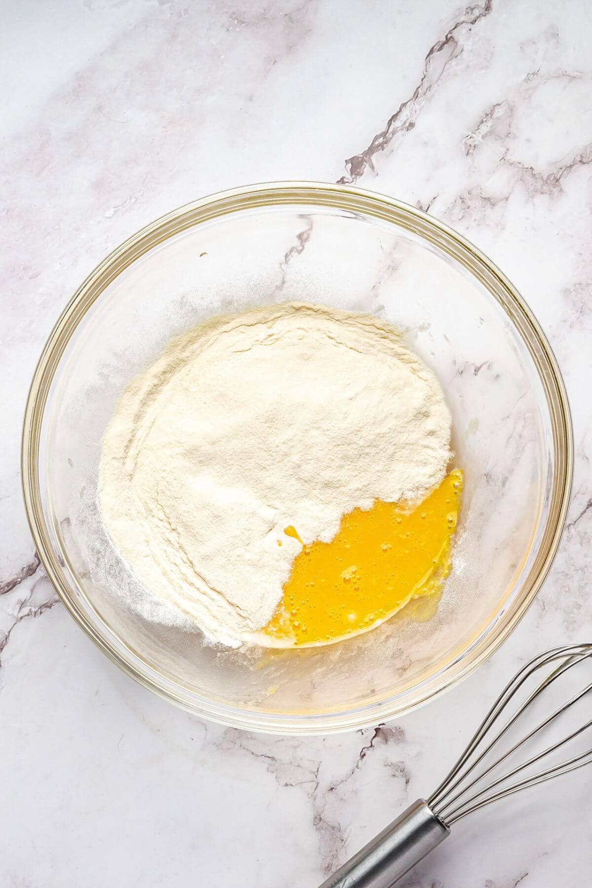 A glass bowl with flour and beaten eggs on a marble surface, next to a metal whisk.