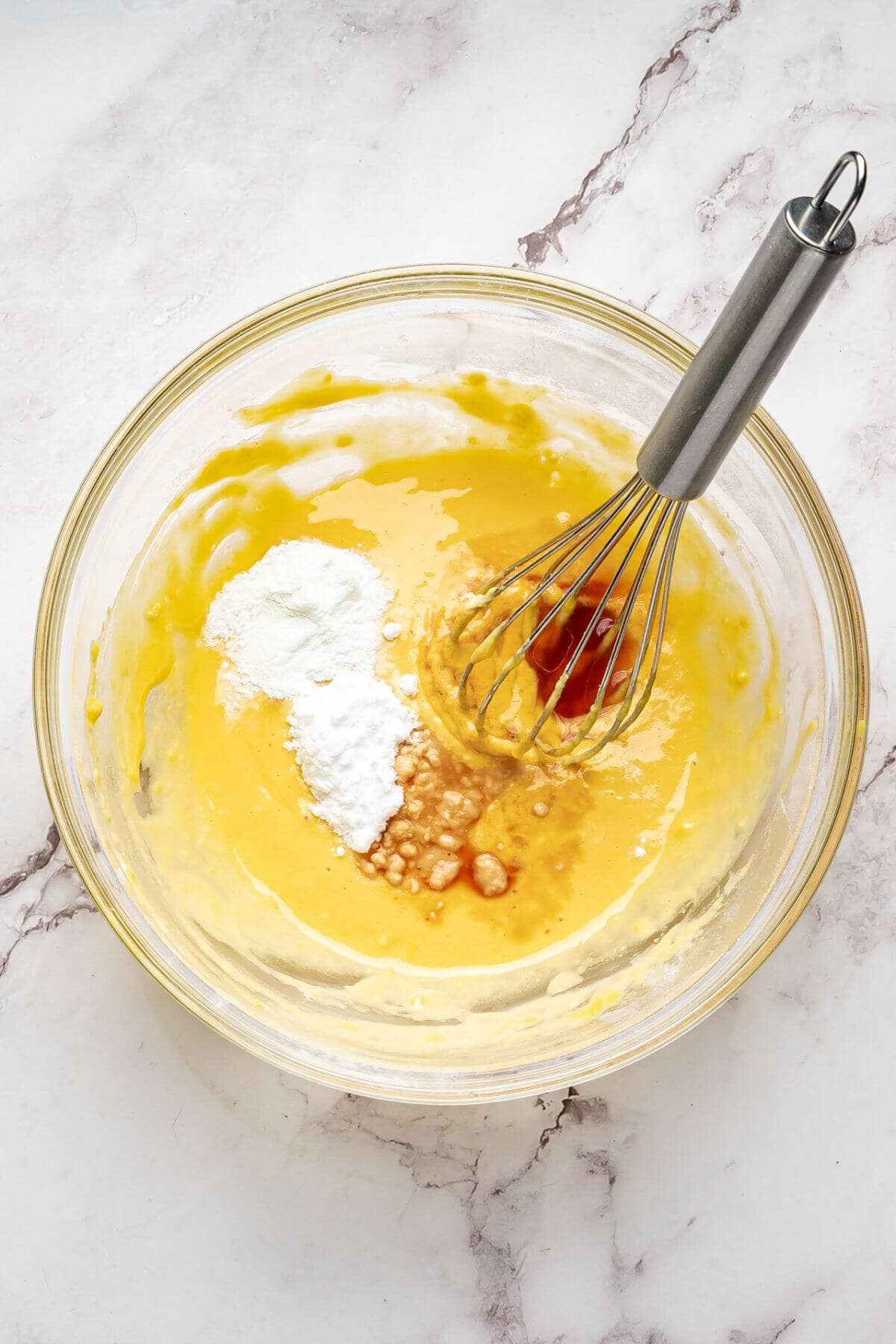 A glass bowl with yellow batter, baking powder, and vanilla extract being mixed with a metal whisk on a white marble surface.
