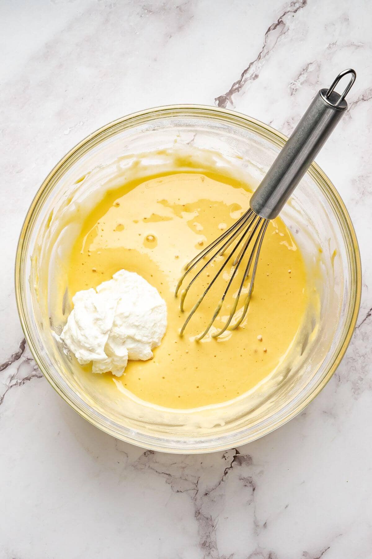 A glass bowl with yellow batter, a dollop of whipped cream, and a metal whisk on a marble surface.