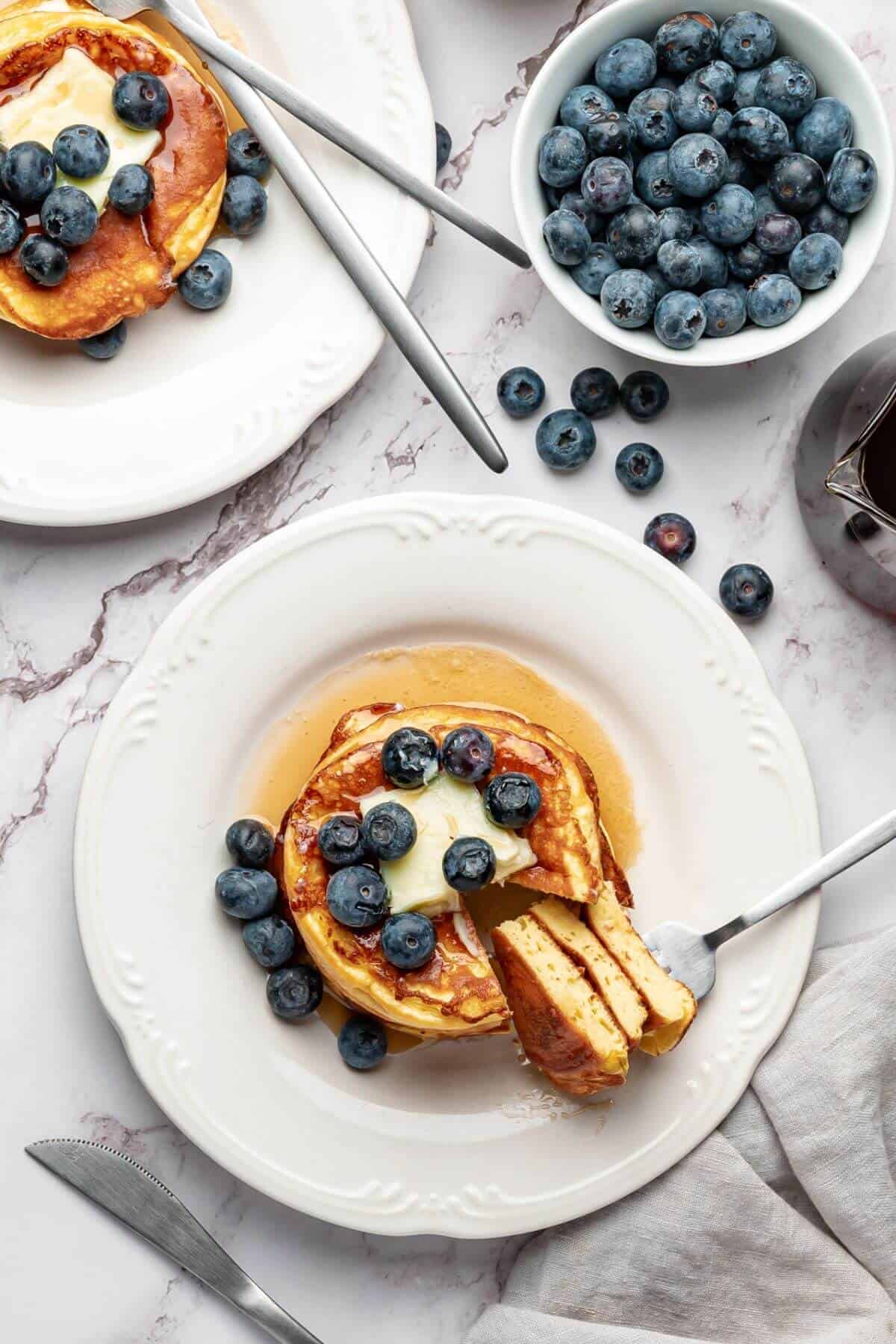 A plate of pancakes topped with blueberries, butter, and syrup, with a fork holding a sliced piece. Nearby are additional blueberries in a bowl and another plate of pancakes.