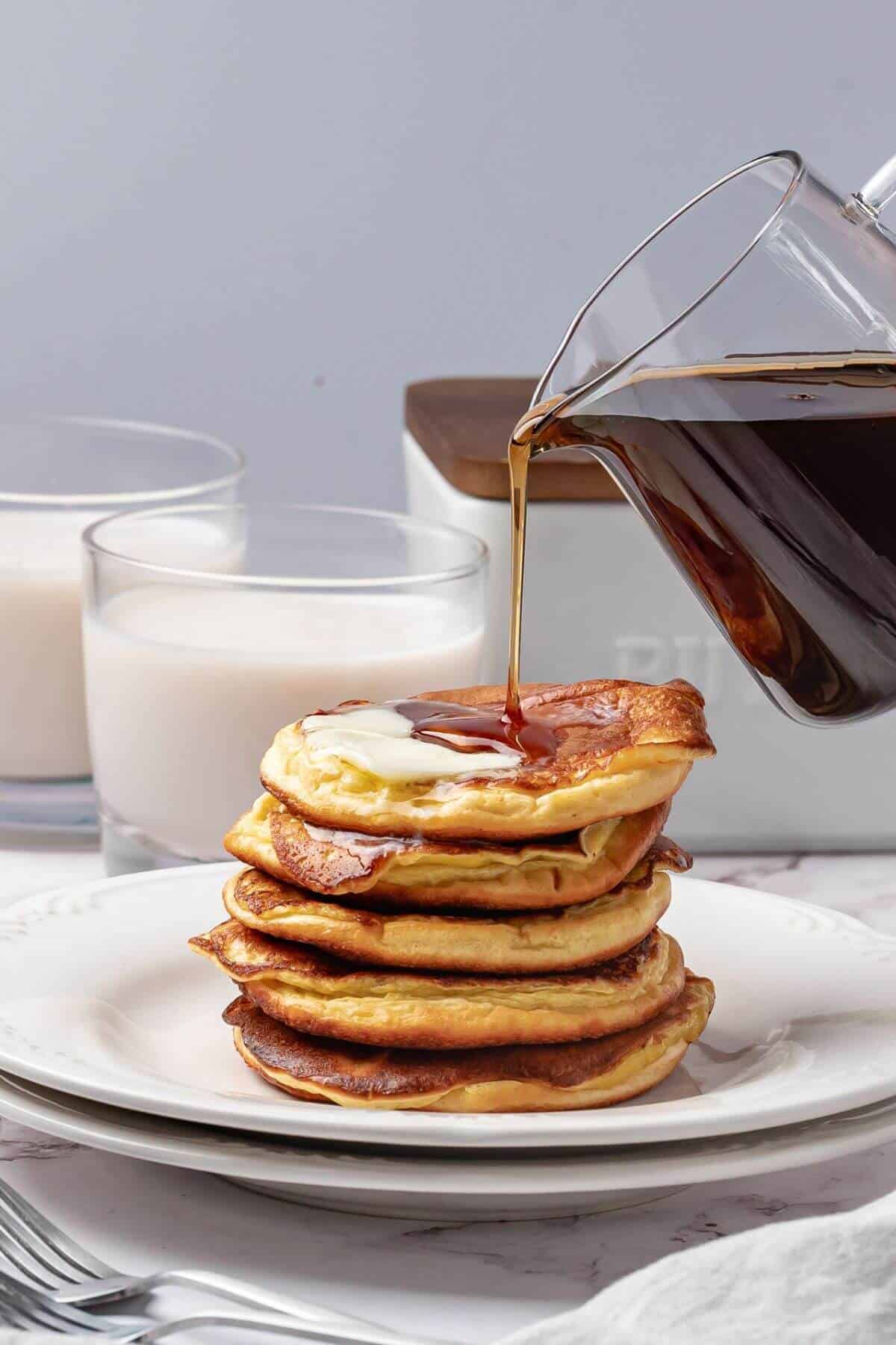 A stack of pancakes topped with butter is being drizzled with syrup, with two glasses of milk in the background on a white table setting.