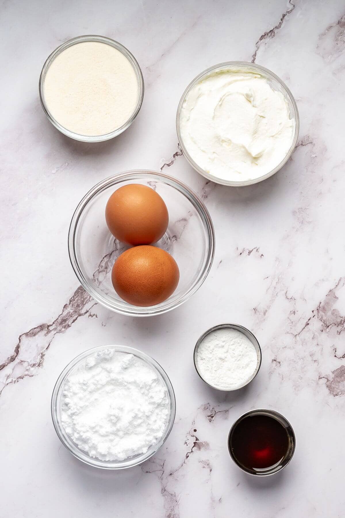 Five bowls and one cup containing granulated sugar, cream cheese, two eggs, powdered sugar, cornstarch, and vanilla extract arranged on a marble surface.