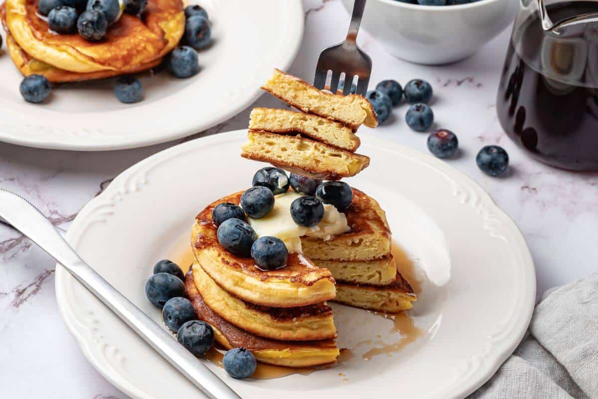 A stack of pancakes topped with butter and blueberries sits on a white plate with syrup; a fork holds a bite of pancakes above the stack.