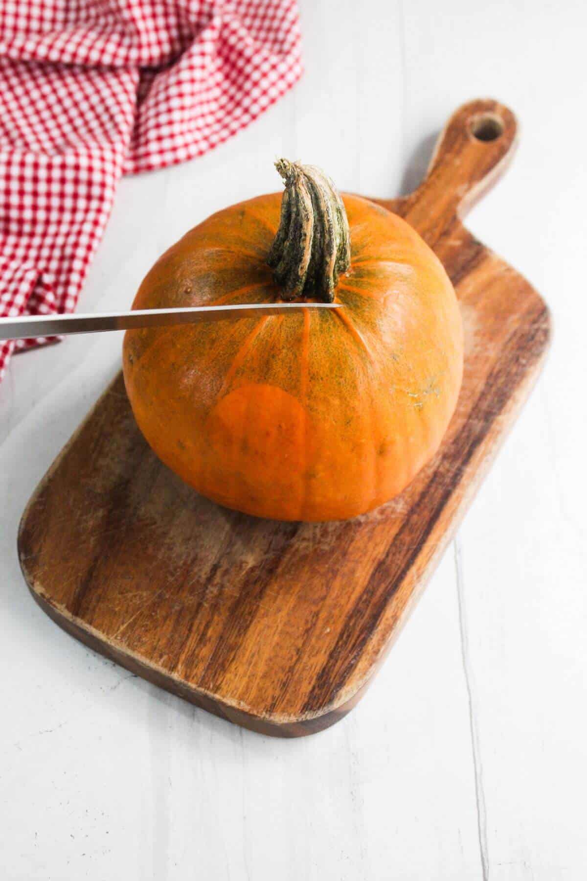 A knife is cutting into the top of a small orange pumpkin placed on a wooden cutting board, with a red checkered cloth nearby.