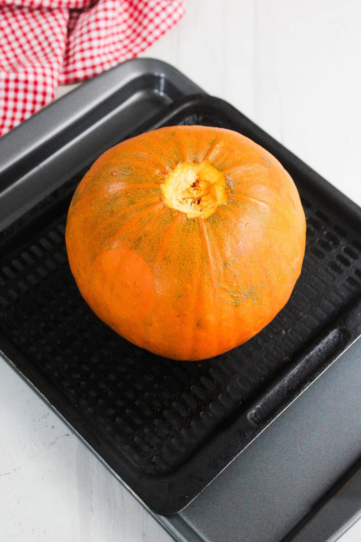 A whole pumpkin sits on a black baking tray, ready for roasting; a red and white checkered cloth is visible in the background.