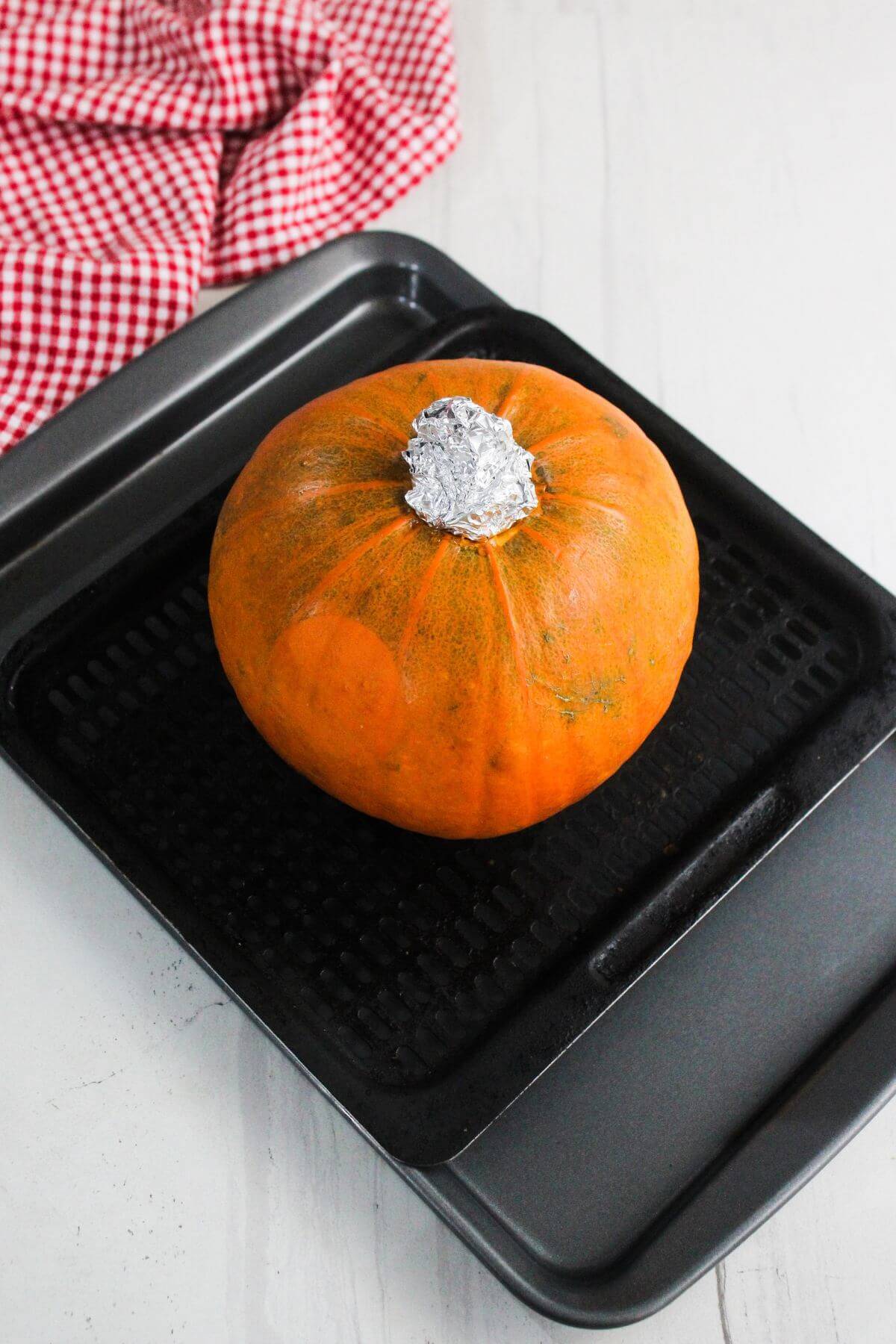 A whole pumpkin with its stem covered in foil sits on a black roasting pan, with a red and white checkered cloth in the background.