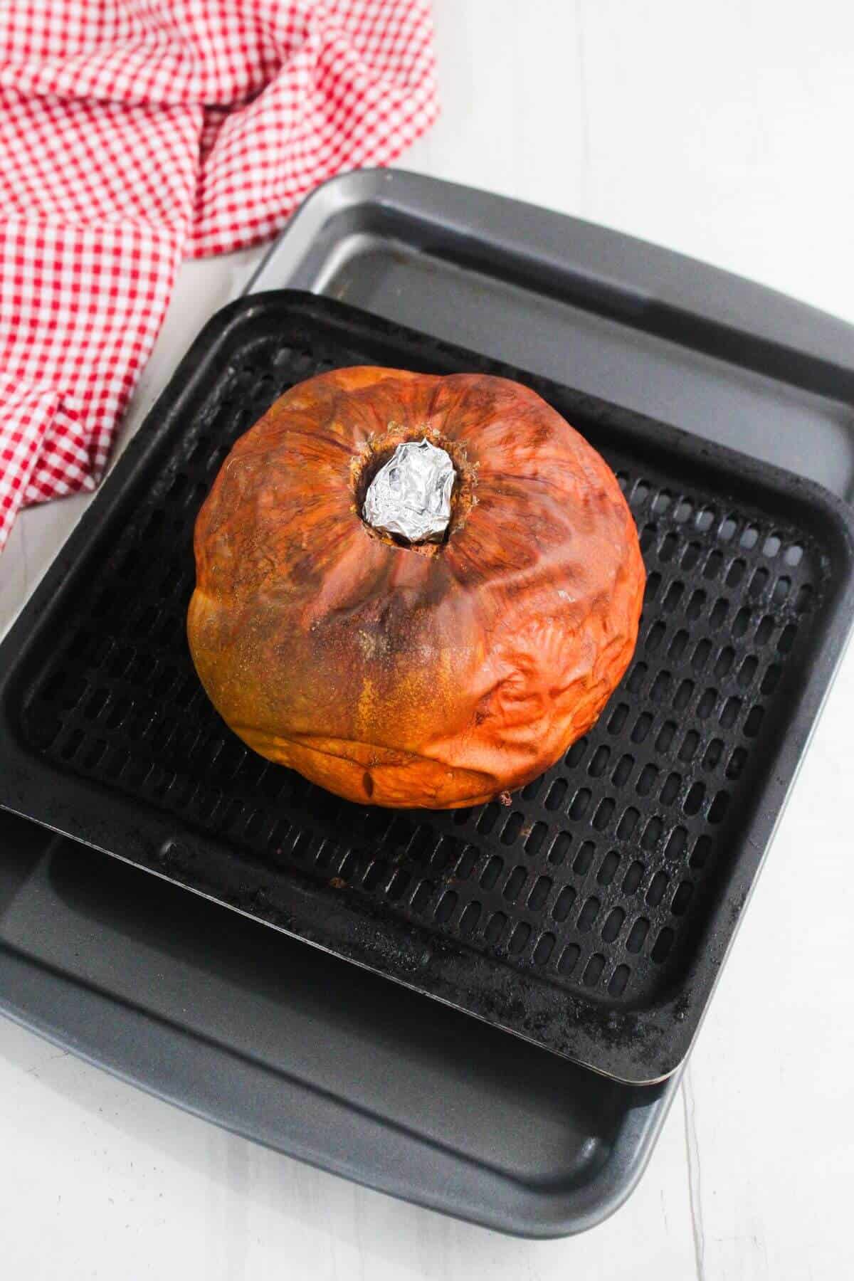 A whole roasted pumpkin with a foil-wrapped stem sits on a black baking tray atop a larger tray, next to a red-and-white checkered cloth.