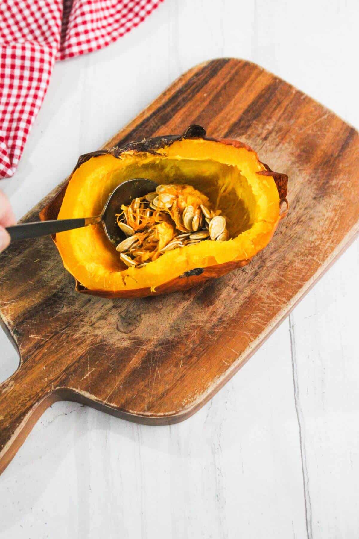 A halved roasted squash with seeds and pulp being scooped out with a metal spoon on a wooden cutting board.