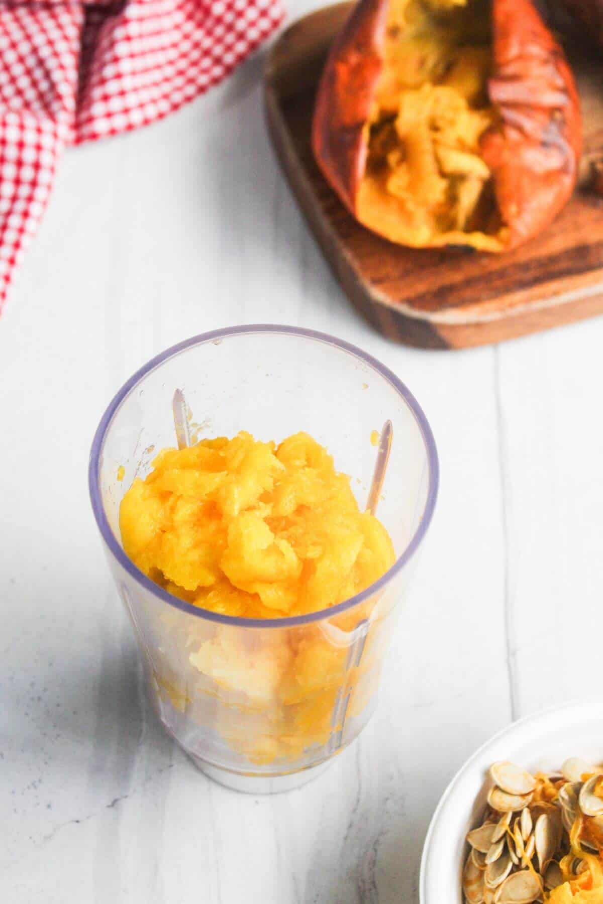 Mashed roasted sweet potato in a clear blender cup on a white surface, with a split sweet potato on a wooden board and some seeds in a small bowl nearby.