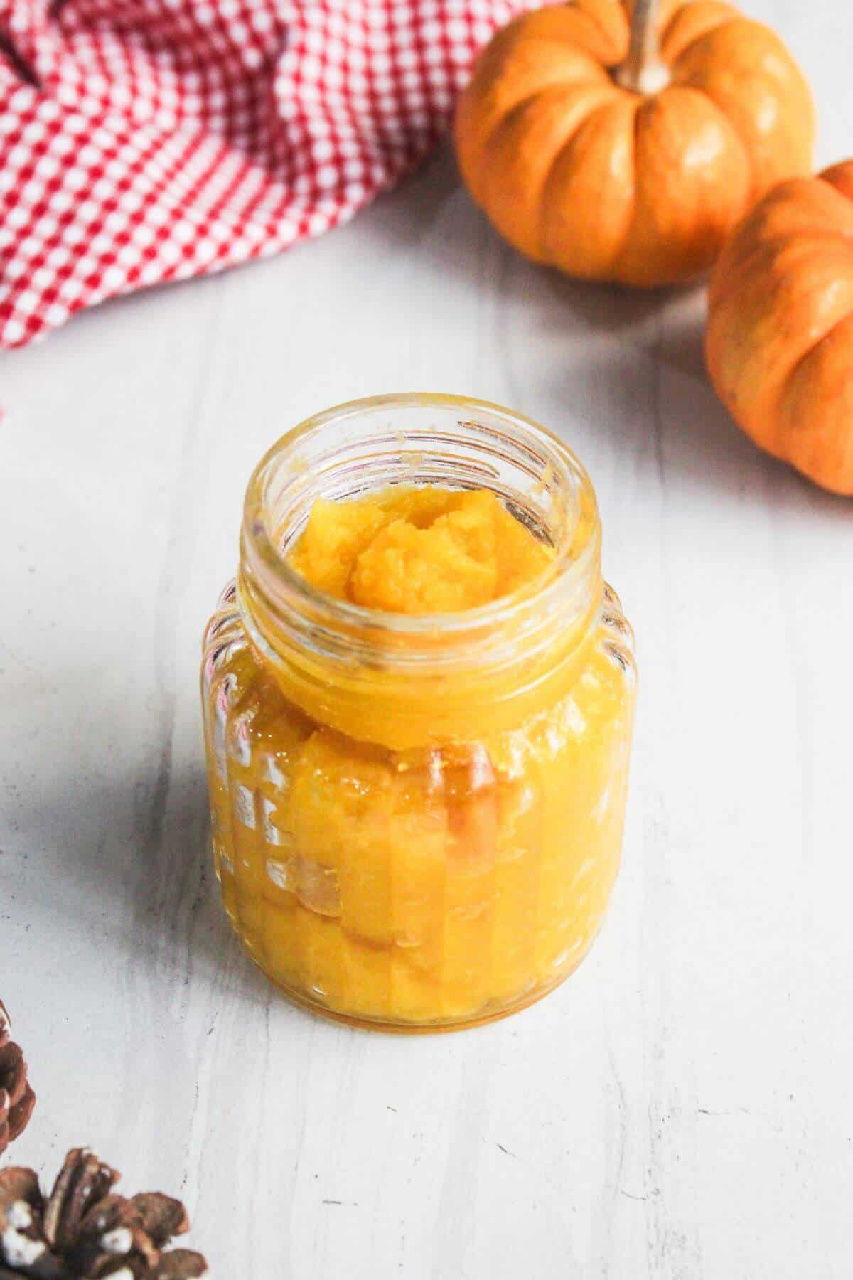 A small glass jar filled with orange pumpkin puree sits on a white surface, with mini pumpkins and a red checkered cloth nearby.
