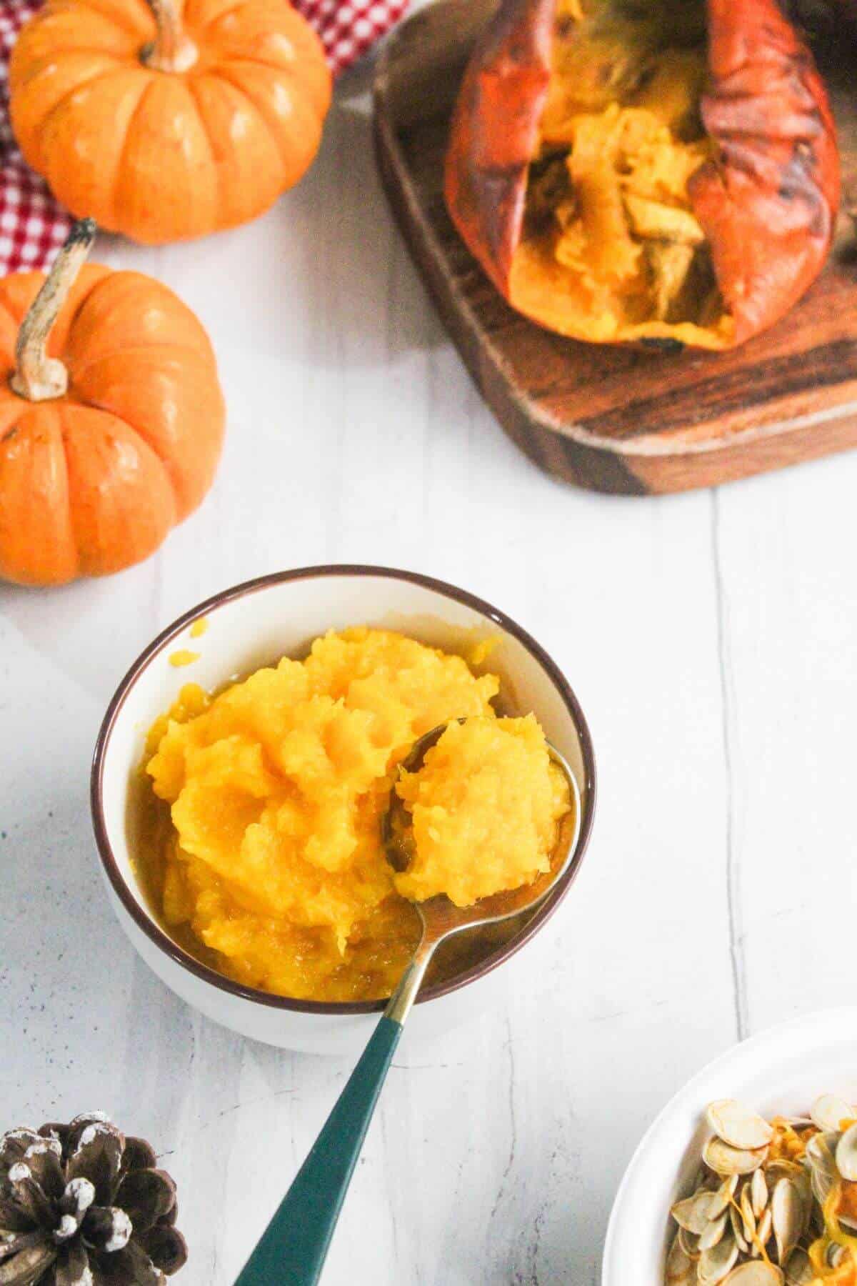 A bowl of pumpkin puree with a spoon, surrounded by whole pumpkins, roasted pumpkin, and pumpkin seeds on a white surface.