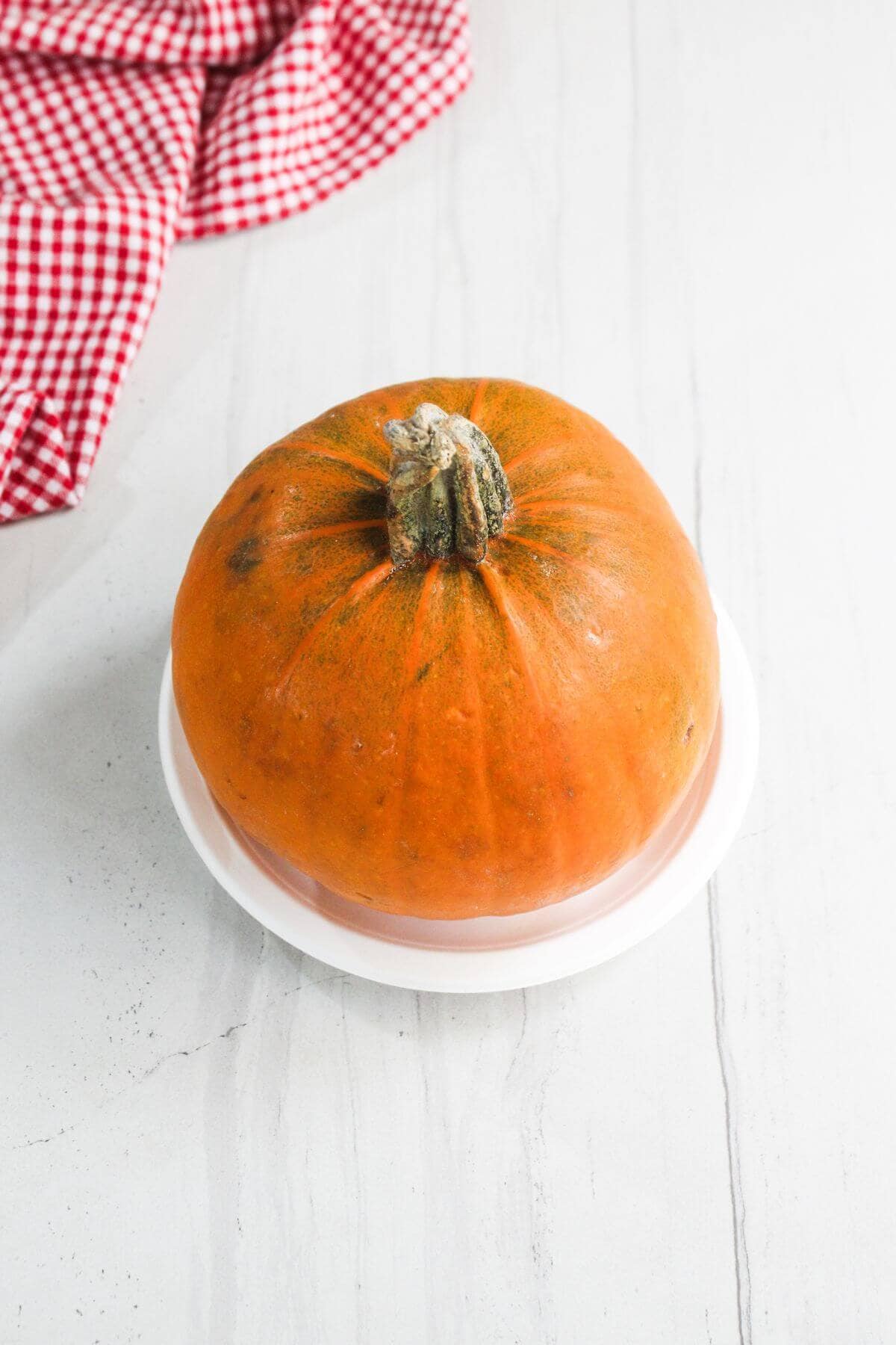 A whole orange pumpkin sits on a white plate atop a light-colored surface, with a red and white checkered cloth in the background.