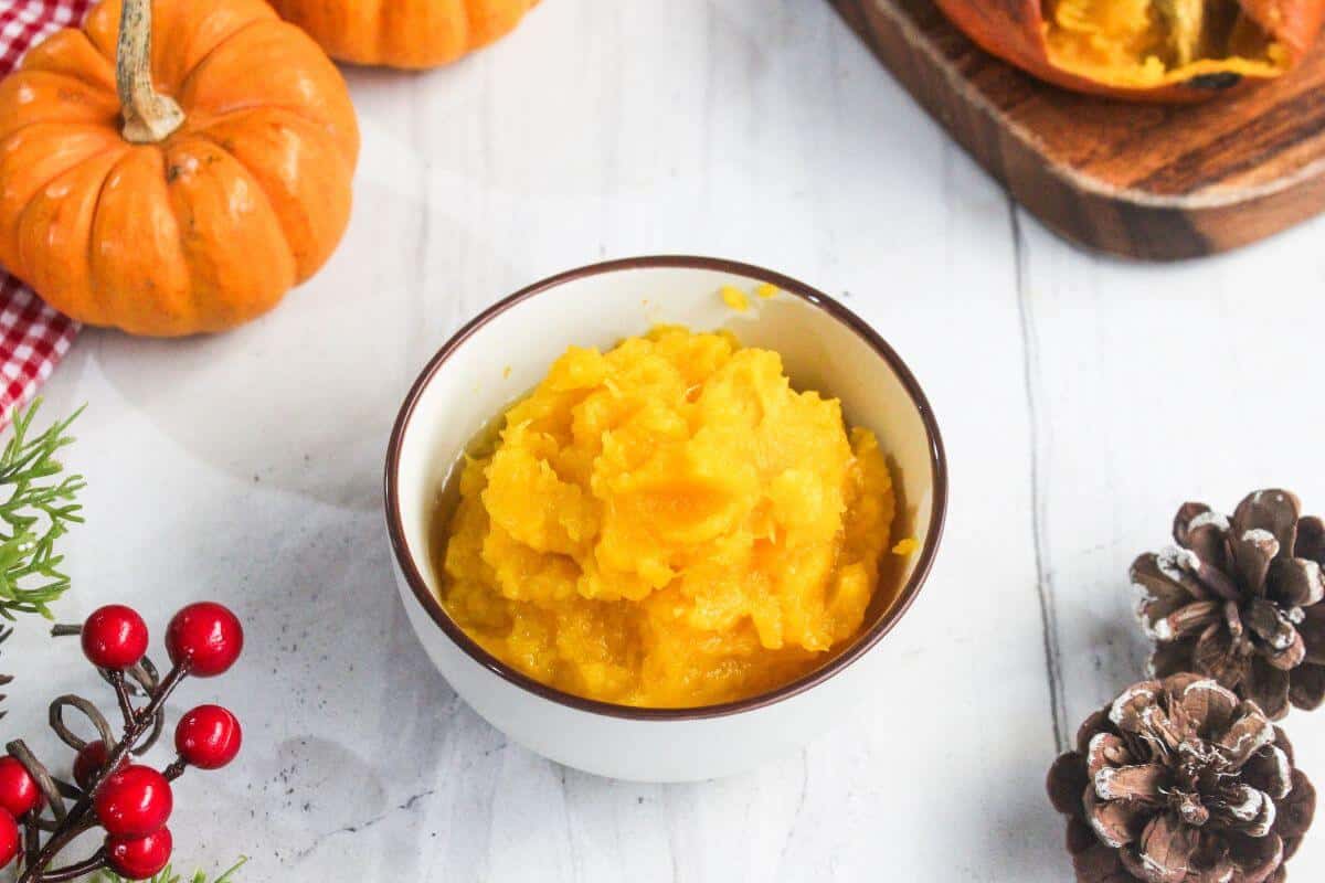 A bowl of mashed pumpkin sits on a white surface, surrounded by small pumpkins, pinecones, red berries, and a wooden cutting board.