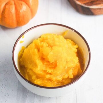 A white bowl filled with smooth pumpkin puree sits on a white surface, with a pumpkin and wooden board partially visible in the background.
