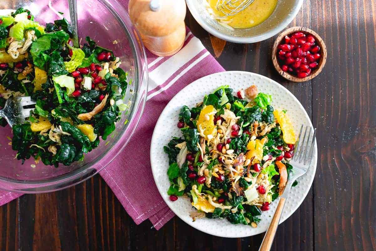 A plate of mixed greens salad with shredded chicken, orange slices, pomegranate seeds, and seeds, next to a bowl of dressing and a pepper grinder on a wooden table.