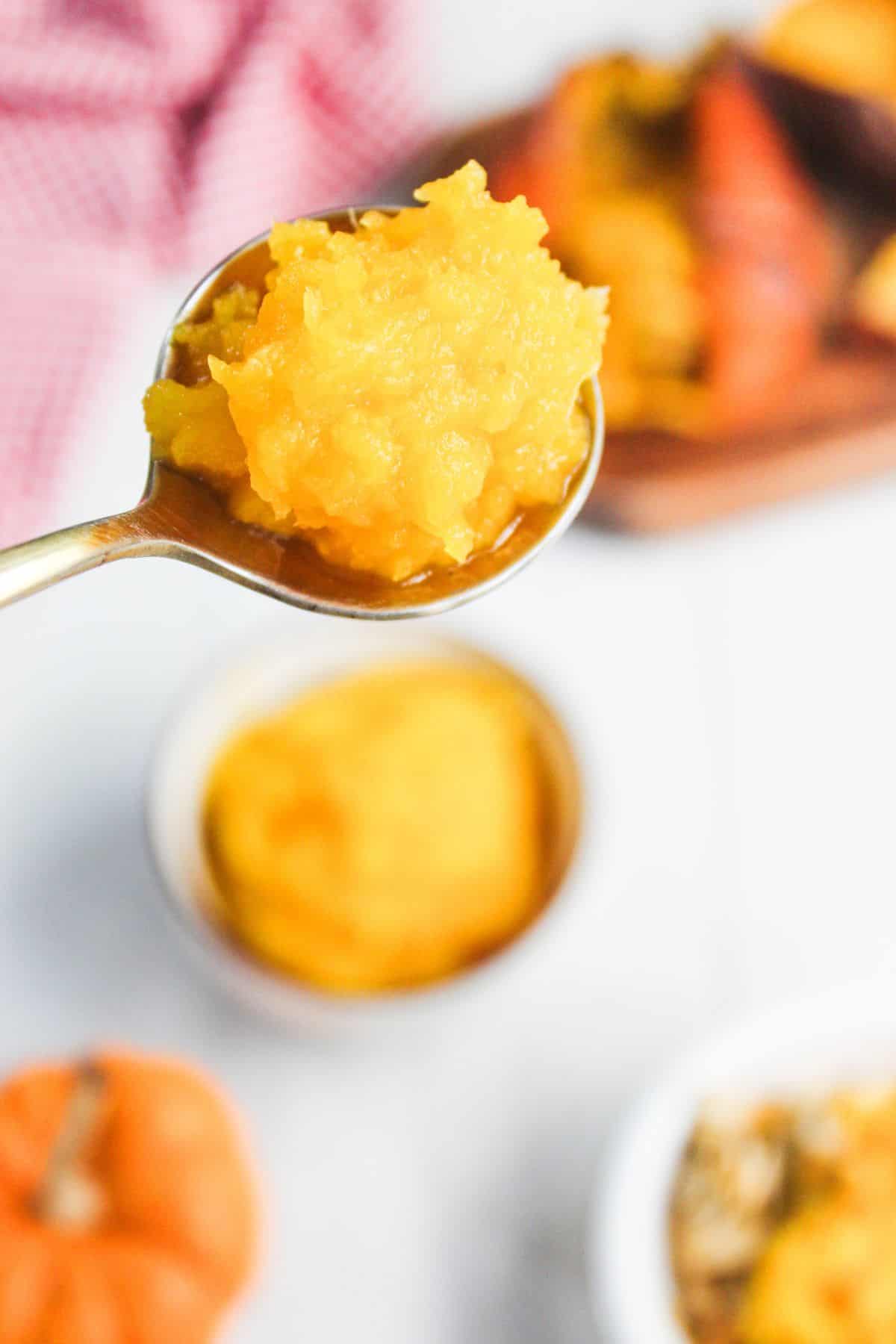 A spoonful of bright orange pumpkin puree held above a bowl, with pumpkins and a red towel in the background.