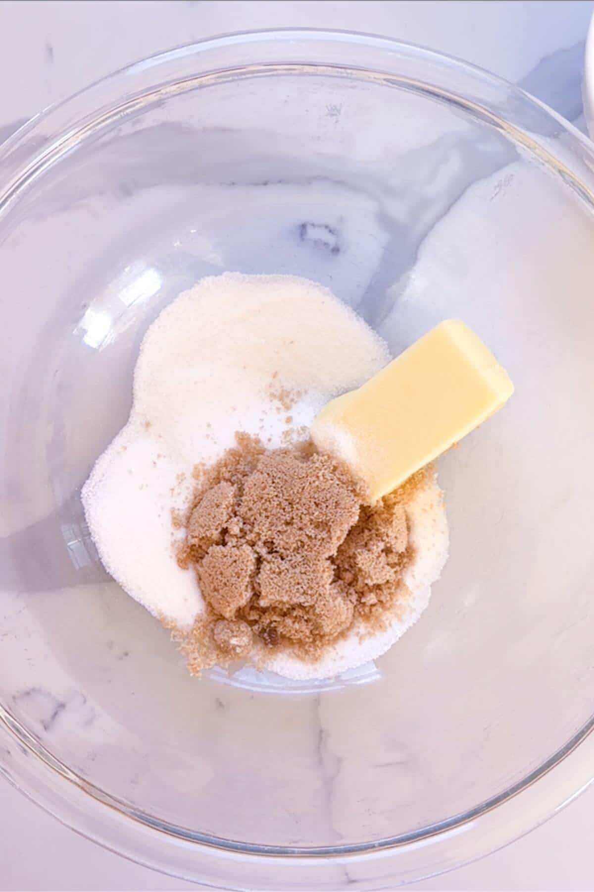 A clear glass bowl containing white granulated sugar, brown sugar, and a stick of butter on a white marble surface.