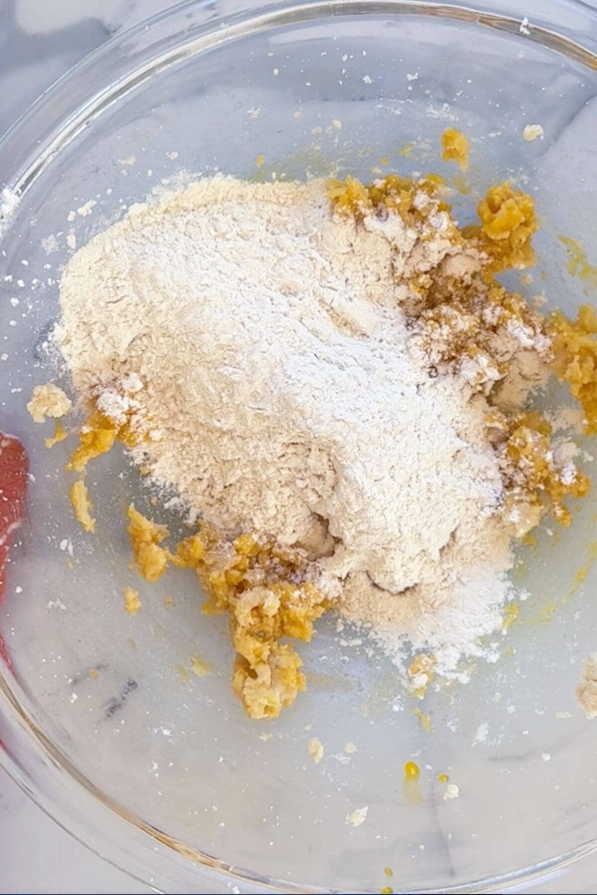 A glass mixing bowl containing partially mixed flour and creamed yellow ingredients, likely for baking, sits on a marble surface.