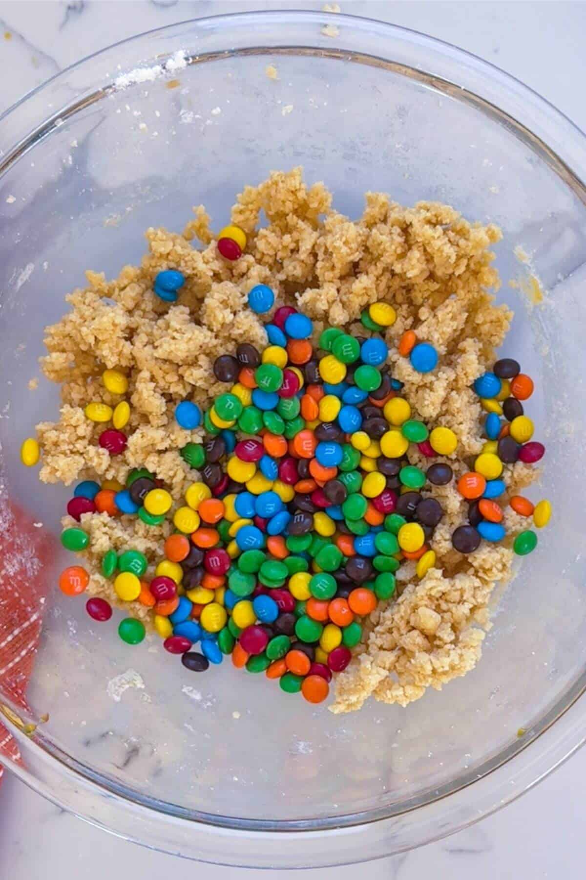 A glass bowl containing cookie dough mixed with colorful candy-coated chocolates on a marble surface.
