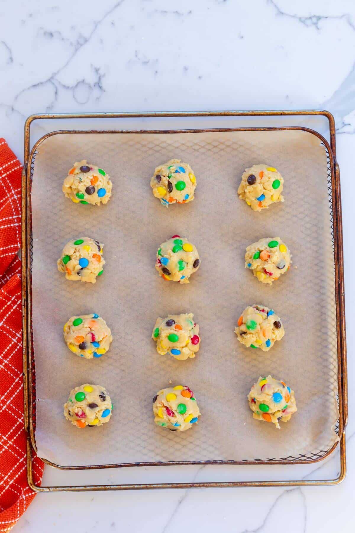 Twelve unbaked cookie dough balls with colorful chocolate candies on a parchment-lined baking sheet, ready to be baked.