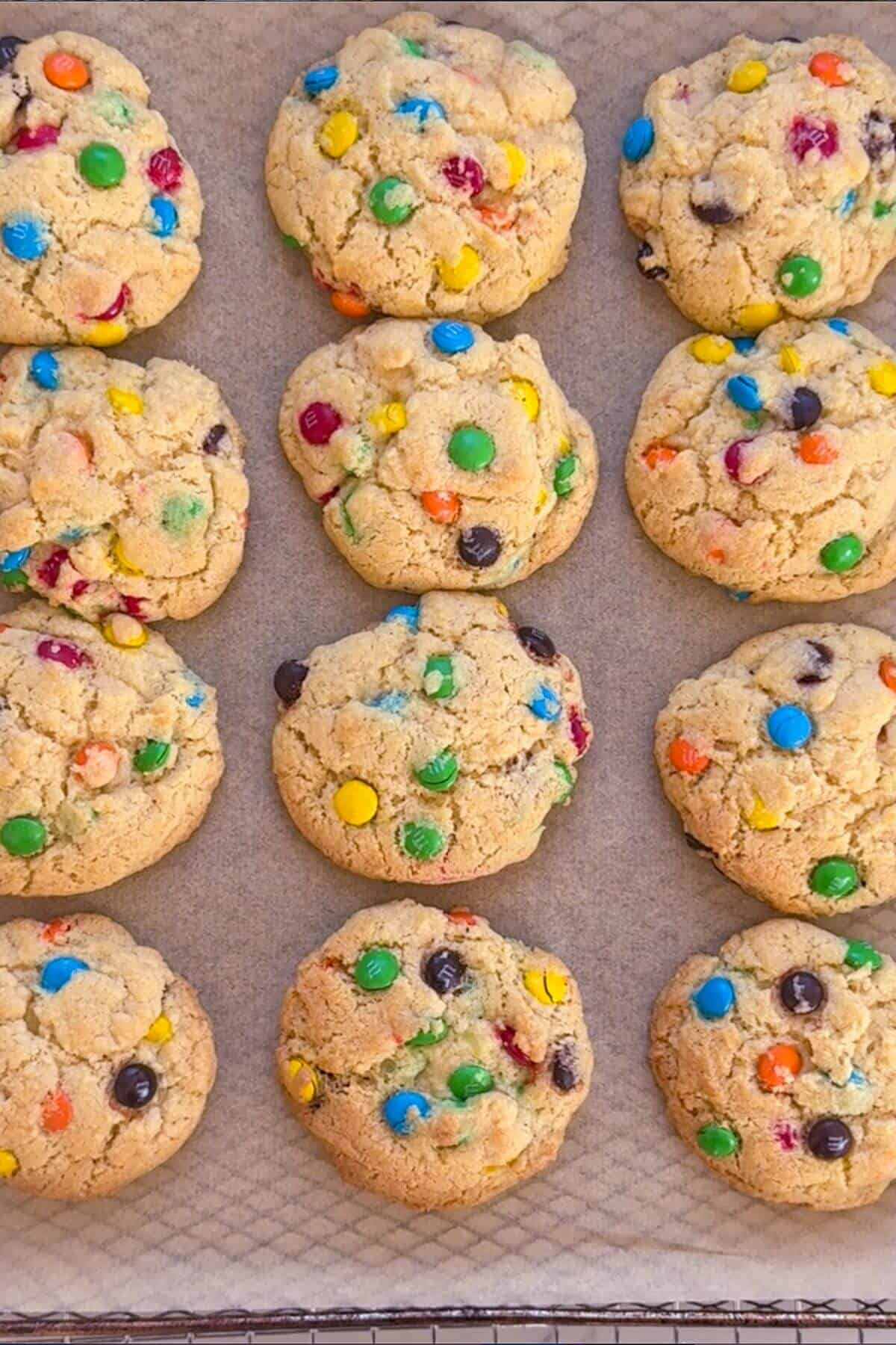A baking tray lined with parchment paper holds twelve freshly baked cookies topped with colorful candy-coated chocolates.